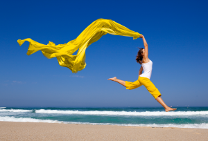 A young woman in yellow pants and a white shirt jumping with a yellow scarf in the wind